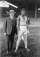 Un homme en costume, portant un chapeau, tient par la hanche un autre homme, habillé en tenue de boxeur. Tous deux se tiennent debout sur la pelouse centrale d'un vélodrome. Derrière eux, la piste et une grande tribune.