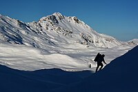 Un excursionist urcă pe un câmp de zăpadă, cu pantele luminate de soare ale unui munte mare și înzăpezit în fundal