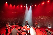 Groupe de rock composé de quatre hommes se produisant sur scène, éclairé par des projecteurs, avec une rangée de fans sombres au premier plan