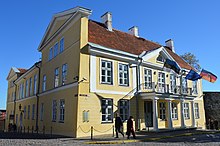 Seitliche Farbfotografie von einem gelben, dreigeschossigen Gebäude mit weißen Umrandungen an Fenstern und Giebeln. Über dem weißen Säuleneingang sind auf dem Balkon Flaggen aus Estland, Europa und Deutschland. Wenige Passanten laufen um das Gebäude herum.