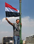 Ung mann med det egyptiske flagget på Qasr al-Nil-brua i Kairo. I bakgrunnen den utbrente bygningen til det tidlegare regjeringspartiet.