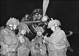 Photographie en noir et blanc à mi-hauteur de 4 militaires debout en treillis et casqués devant un avion à hélice. Ils regardent leur montre.