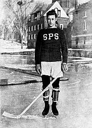 Black and white photo of a young boy posing on an ice rink. He is wearing skates and gloves, while holding a hockey stick. He's wearing a sweater with the letters "SPS" on it