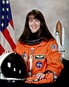 image of Janet Kavandi in 2001, posing in her orange NASA uniform with helmet in front of her, and U.S. flag and an upright Space Shuttle model to each side of her in the background