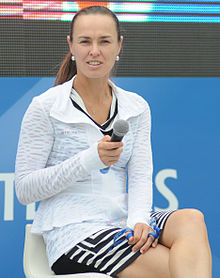 Photo couleur d'une femme assise sur une chaise, de trois-quart face, sur un court de tennis, un micro à la main. Elle porte un polo blanc par dessus une robe à motifs noir et blanc