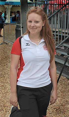 Photo d’une jeune femme, rousse, portant un polo de sport blanc aux manches rouges et des lunettes à soleil dans les cheveux