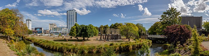 Detrás de un pequeño río serpenteante y un parque arbolado podemos ver algunas construcciones.