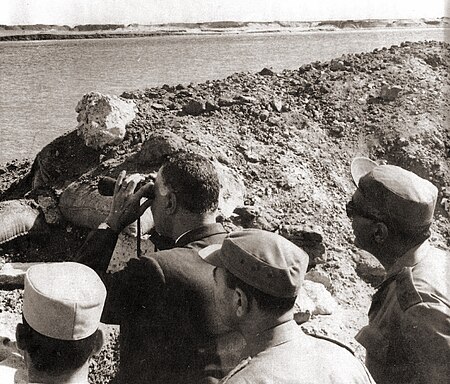 A man wearing suit peering out across a body of water with binoculars from an opening in dirt mound. Behind him are three men in military uniform