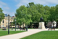 An equestrian statue stands at the centre of a grass-covered square, with several paths leading towards the centre from the corners and sides in a star-shape. People sit on benches or walk on the paths.