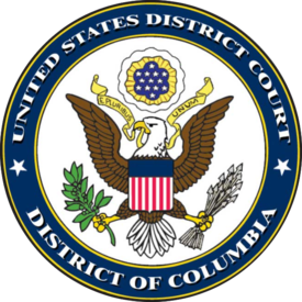 A bald eagle is holding a yellow ribbon in its mouth, which reads "E Pluribus Unum". The eagle also is holding an olive branch in one talon, and arrows in the other. Around the bird is a blue border that reads, "United States District Court: District of Columbia".