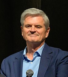 head and shoulders crop of Case wearing light blue shirt and navy suit jacket, holding a microphone and facing audience