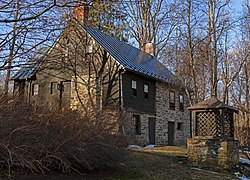 A two-and-a-half-story stone house with a gabled roof and brick chimneys built into a hillside, seen from a three-quarters angle. It has brown wooden insets in the gable fields on the side. There are small patches of snow in front and the surrounding trees are bare, lit from the left by a low sun. In front left is a large stone structure topped by latticed square-shaped woodwork with a gabled roof.