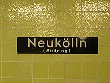 A dark blue sign on a yellow tiled station wall, bearing the name 'Neukölln (Südring)'