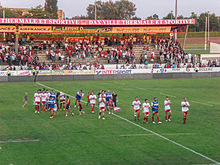 Rugby oyuncuları çimenlerde yürür;  Arka planda taraftarlar bir stanttan alkışlıyor.