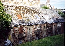 dilapidated single-storey brick block of four cells with wooden doors under a slate roof with moss growth and stone buildings behind