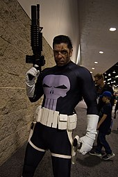 photo en couleur d'un homme déguisé en Punisher (costume gris avec un crâne stylisé sur le torse) dressant de la main droite une fausse mitraillette