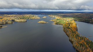 Photographie aérienne d'une partie d'un lac dans un paysage plat, nuagé, couvert de forêts.