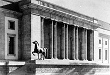 Seitliche Schwarzweißfotografie eines Gebäudes mit einem mittleren Säulenvorhof. An beiden Seiten sind ein langes Fenster und darüber quadratische Fenster zu sehen. Links führen Stufen zum Eingang und werden von einer Mauer begrenzt, auf der links eine Pferdeskulptur trabt.