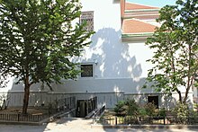 A large white building with 2 trees in front and a lower entrance