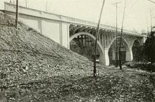 Vista em três quartos em preto e branco logo acima do nível da água de uma ponte em arco de spandrel aberto de concreto armado que atravessa um rio.