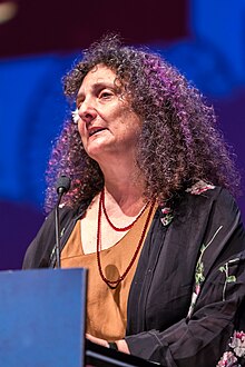 middle-aged white woman standing at a podium, speaking into microphone