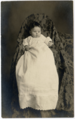 Baby in einem langen weißen Kleid auf einem scheinbar mit Stoff umhüllten Stuhl.