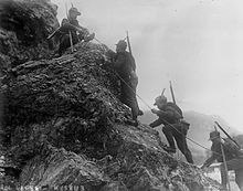 Čtyři italští pěšáci šplhající s pomocí lana v hornatém terénu.