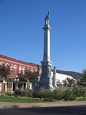A stone monument about three stories tall rises from a park-like area along a city street. The monument features stone replicas of uniformed soldiers.