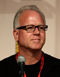 A man with white hair and glasses sits behind a microphone.