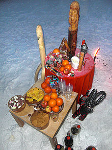 Two tables located in the snow. On the tables are various fruits and wooden quasi-anthropomorphic statues.