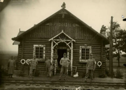 Six Austrian-Hungarian soldiers and their duck stand in front of a wooden station building beside the narrow gauge railway track