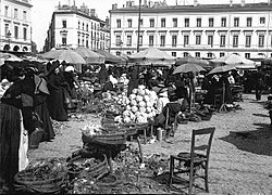 Photographie du marché du Capitole prise depuis le centre, en direction du nord-ouest. Au premier plan paniers de fleurs, chaises ; au second plan étals sous parosol, personnages ; en arrière-plan façades sur la place.