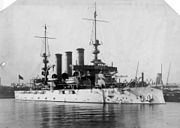 A white battleship with three smokestacks and two tall masts sitting in port.