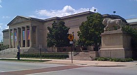 Photo d'un grand bâtiment avec des piliers de style grec, des arbres et une statue de lion à proximité.