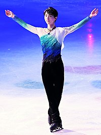 2017年世界選手権の表彰式にて観客の声援に応える羽生結弦。スケートリンクの上で足をクロスして腕を広げて微笑んでいる。