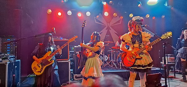 During live performances, referred to as "servings", band members wear elaborate maid costumes (L-R: Misa, Kanami Tōno, Miku Kobato; Boston, 2022)