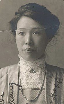 Yüksek dantel yakalı bluz, boncuklar ve ceket giyen genç bir Asyalı kadın; koyu saçları kabarık. Alt kenarından fotoğrafın üzerinden geçen bir el yazısı var.
