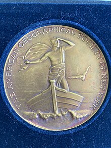 Image of bronze circular medal in dark blue velvet. The medal includes the text "The American Geographical Society of New York" in all capital letters around the edge of the medal. The center image on the medal is a man standing on a boat looking to his left with a bird on the left.
