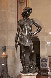 Fotografía de la estatua de bronce de un joven vestido con ropa ligera que sostiene una espada corta en la mano derecha con la mano izquierda en la cadera.  De pie en una postura triunfante y relajada entre sus pies se coloca sobre una cabeza cortada.