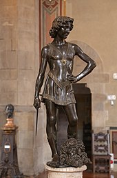 Fotografia della statua in bronzo di un giovane vestito con abiti leggeri che tiene una spada corta nella mano destra con la mano sinistra sul fianco.  In piedi in una postura trionfante e rilassata tra i suoi piedi è posto su una testa mozzata.