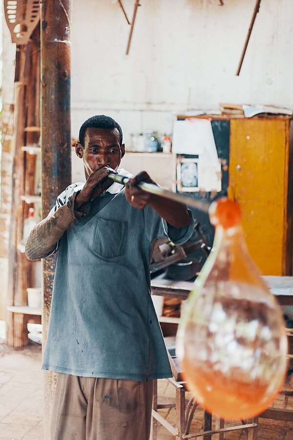 A glassworker blows air into the glass, creating a cavity inside