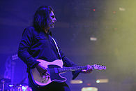 A man plays guitar on a major stage set wearing a black suit and a mask that resembles black and white makeup.