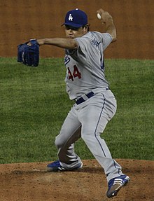 Ein Mann, der eine graue Baseballuniform und eine blaue Baseballkappe trägt, wirft einen Baseball mit seiner rechten Hand.