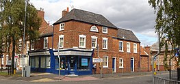 The corner of a terraced suburban street. The lower storey is a corner shop, advertising as a chiropractic clinic. The building is two storeys high, with some parts three storeys high.