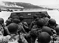 American assault troops in a landing craft huddle behind the protective front of the craft as it nears a beachhead, on the Northern Coast of France. Smoke in the background is Naval gunfire supporting the land.