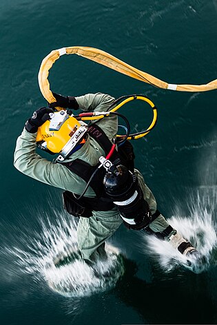 Дайвер, снабженный надводным оборудованием ВМС США, одетый в легкий шлем и держащий шлангокабель на уровне головы, входит в воду, прыгнув в воду. Вид с палубы, с которой прыгнул дайвер, и показывает спину дайвера в виде ласт первый контакт с водой