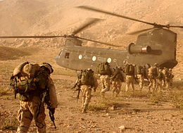 A line of soldiers carrying equipment on their backs walking toward a transport helicopter in desert terrain