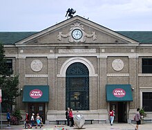 The central three bays of a large brick train station. A pilaster with a large clock tops the three bays. The central bay has an ornamental grille; the flanking bays have statues of Mercury and entrance doors.