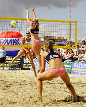 Figures féminines sur le sable.  Deux sont du côté proche d'un filet et un de l'autre côté.  Deux sautent.  À droite, un public.