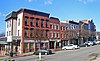 A row of two-story commercial brick buildings, some painted, on a street curving away from the viewer to the right. In front of them are some short bare trees and diagonally parked cars.